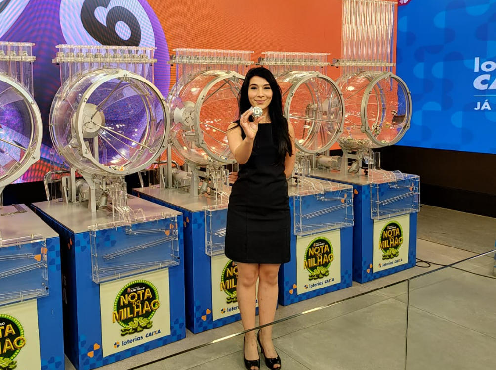 A imagem mostra uma mulher em pé, ao centro, vestida com um vestido preto e sapatos de salto. Ela está sorrindo e segurando uma bola branca com o número “6” em frente ao corpo, voltada para a câmera. Atrás dela há cinco globos transparentes de sorteio, alinhados lado a lado, usados em loterias, com estruturas metálicas e bases azuis. Em cada base há o logotipo da campanha “Nota do Milhão”, com o selo das Loterias CAIXA. O fundo é composto por painéis iluminados com cores vivas — à esquerda, tons de roxo e laranja, e à direita, um grande painel azul com o texto parcialmente visível “Loterias CAIXA”. O ambiente é um estúdio de gravação ou local de sorteio oficial, bem iluminado e organizado.