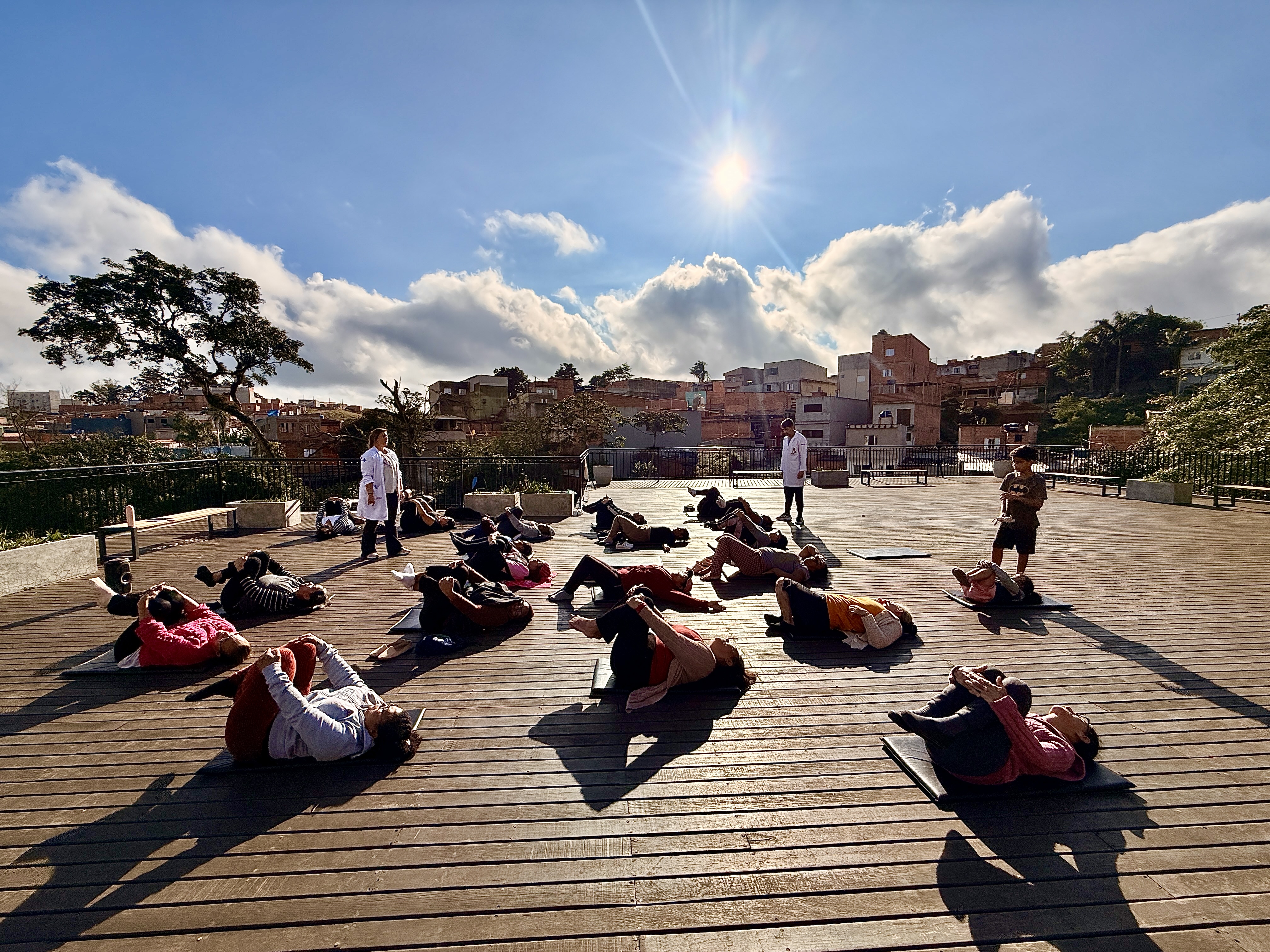 Fotografia colorida feita ao ar livre em um amplo deck de madeira. Cerca de vinte pessoas participam de uma atividade de ioga, deitadas sobre colchonetes e realizando alongamentos em diferentes posições. Duas profissionais de jaleco branco acompanham o grupo. Ao fundo, vê-se uma comunidade urbana em uma área elevada, cercada por árvores e vegetação. O céu azul ocupa grande parte da imagem, com nuvens brancas volumosas e o sol brilhando intensamente, projetando longas sombras sobre o deck. A cena transmite sensação de tranquilidade, bem-estar e integração entre saúde, natureza e território.