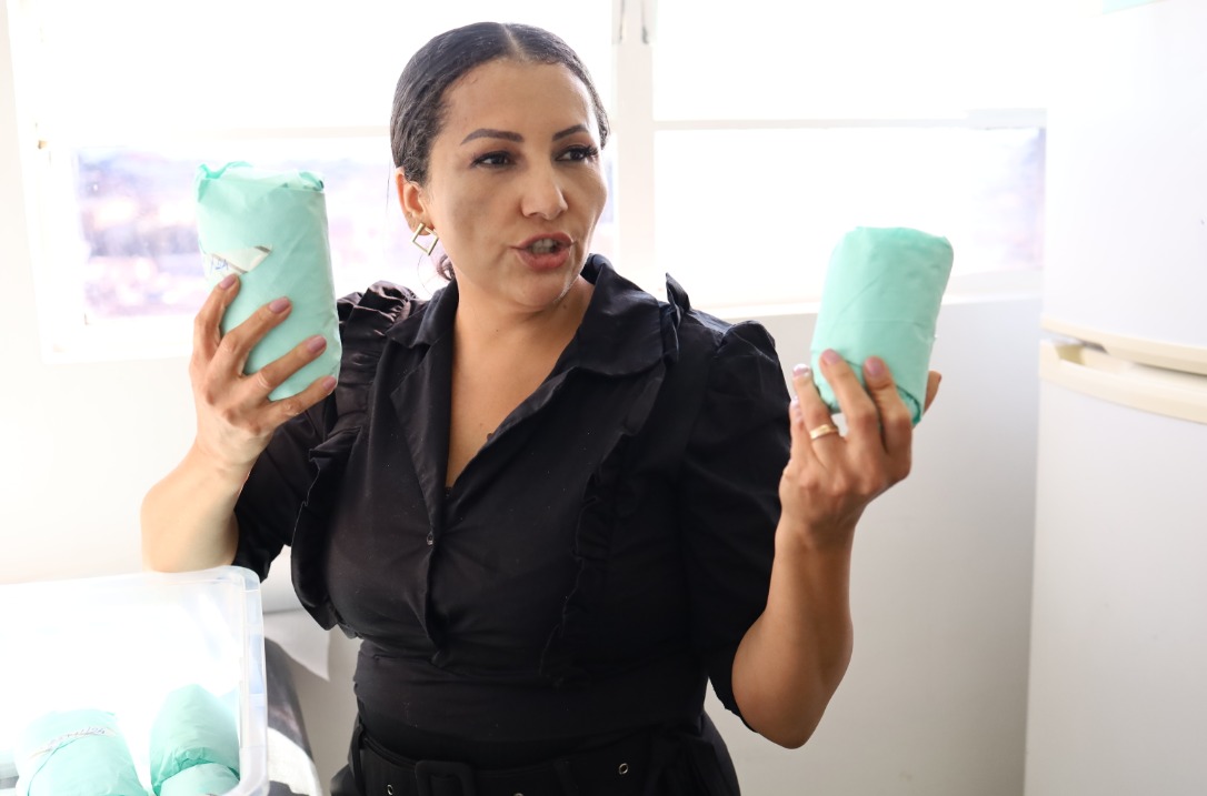 Uma mulher de pele clara e cabelos escuros presos em um coque está no centro da imagem, segurando pacotes cilíndricos envoltos em papel cirúrgico verde (papel grau cirúrgico) são, muito provavelmente, frascos de vidro para armazenamento de leite que passaram pelo processo de esterilização em autoclave.