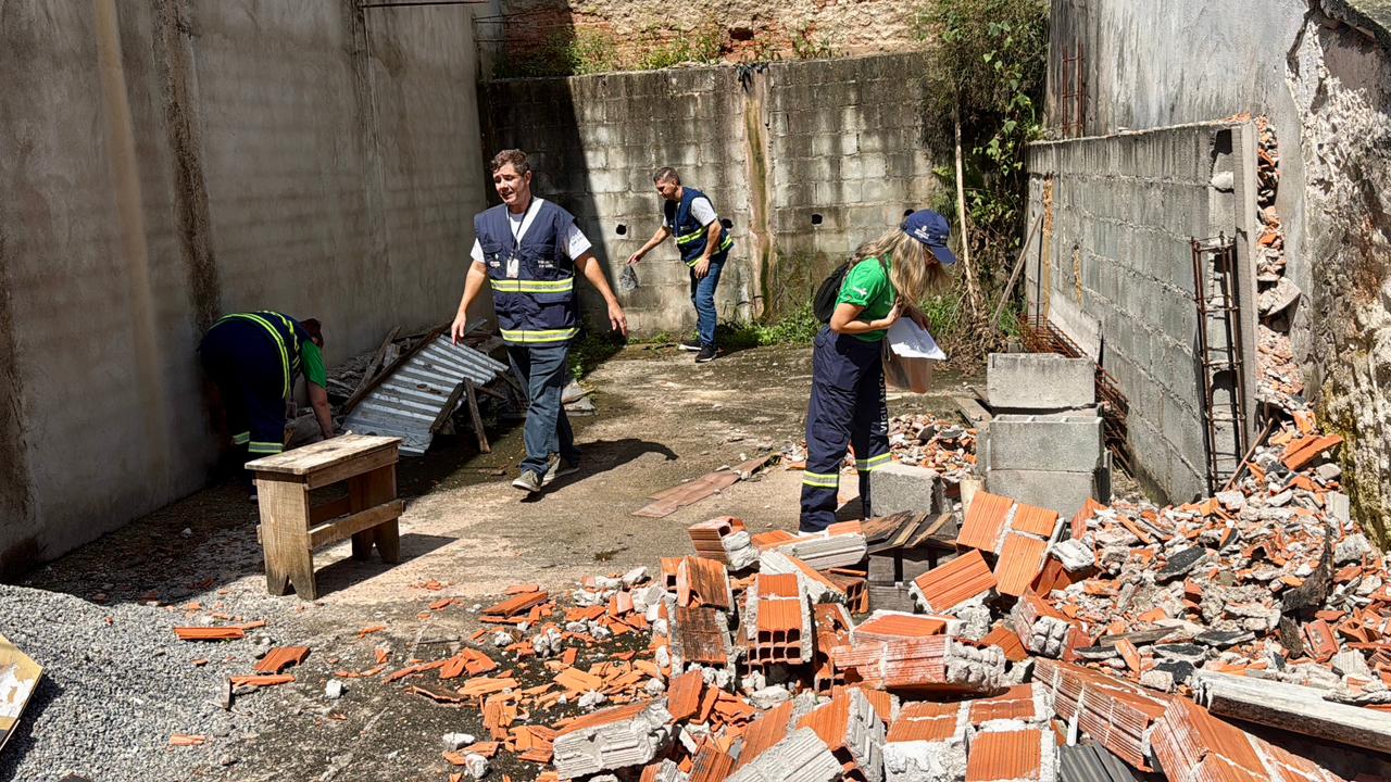 A imagem mostra uma equipe de agentes de saúde realizando uma ação de combate à dengue em um terreno com entulho e materiais de construção. Quatro profissionais, usando uniformes com faixas refletivas, vistoriam o local em busca de possíveis criadouros do mosquito Aedes aegypti. No primeiro plano, há tijolos quebrados e acúmulo de resíduos, enquanto ao fundo é possível ver paredes inacabadas e áreas com vegetação. Um dos agentes recolhe materiais, outro observa o ambiente, e os demais realizam inspeção minuciosa no espaço.