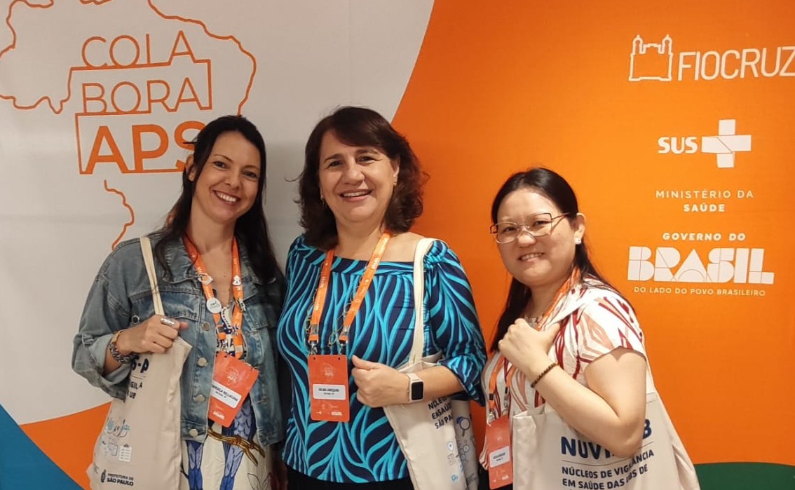 Três mulheres posam sorrindo em frente ao painel oficial do III Encontro Nacional da Rede ColaboraAPS, realizado em Brasília. Ao centro está Selma Anequini Costa, assessora técnica da Coordenadoria da Atenção Básica (CAB) da Secretaria Municipal da Saúde de São Paulo e responsável pela implementação dos núcleos. As participantes usam crachás do evento e carregam bolsas institucionais. Ao fundo, aparecem as marcas da Fiocruz, do SUS e do Ministério da Saúde.