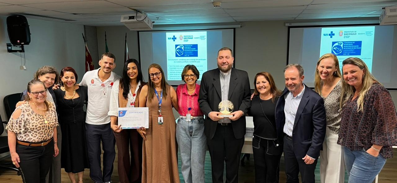 A imagem mostra um grupo de cerca de dez pessoas reunidas em uma sala de eventos ou auditório, posando para uma foto após uma cerimônia de reconhecimento ou premiação. No centro da cena, uma mulher segura um certificado enquanto um homem vestido com traje clerical preto segura um troféu em formato circular dourado.  Os participantes estão sorrindo e posicionados lado a lado, em clima de celebração e confraternização. Ao fundo, dois telões exibem uma apresentação institucional da Secretaria Municipal da Saúde de São Paulo, com o logotipo do SUS e informações relacionadas a um congresso ou encontro na área da saúde.