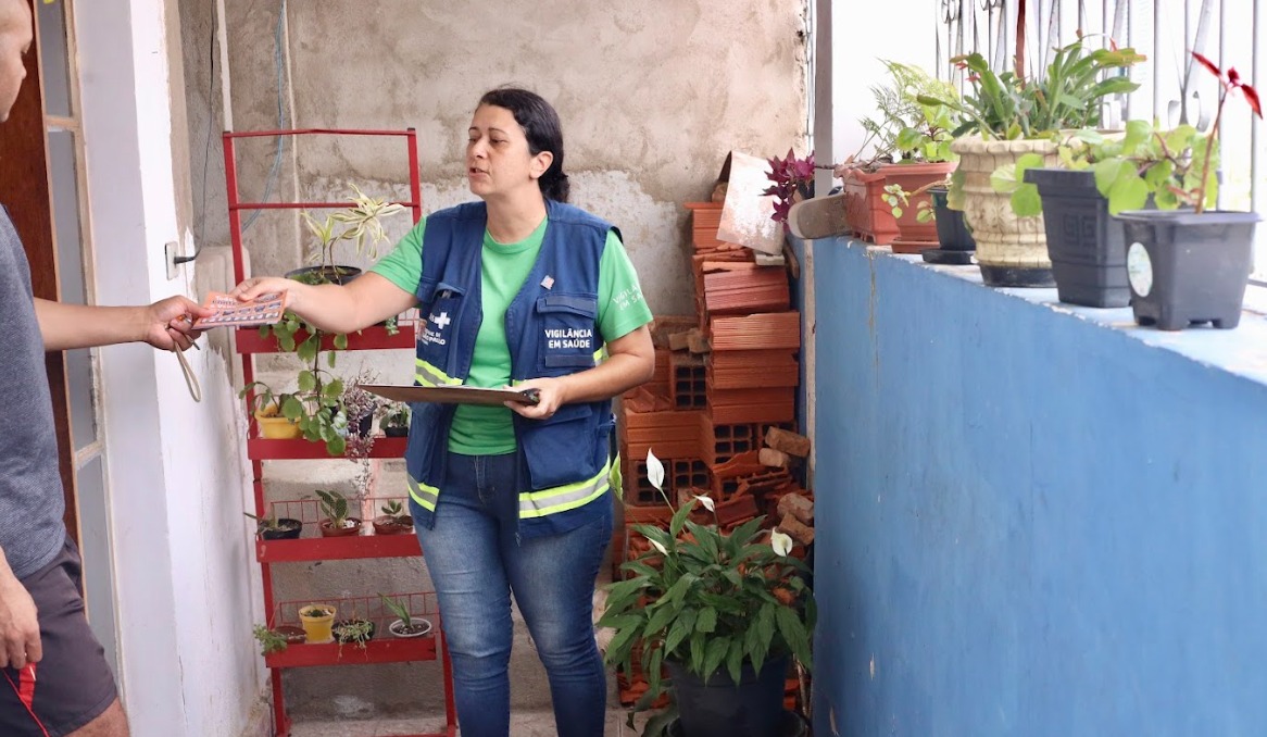 A imagem mostra uma agente de saúde em visita domiciliar, conversando com um morador em uma área externa de uma casa. Ela veste camiseta verde e colete azul com identificação de “Vigilância em Saúde”, segura uma prancheta e entrega um material informativo ao homem, que aparece parcialmente à esquerda.  O ambiente é simples, com paredes sem acabamento completo e um pequeno espaço com plantas em vasos — alguns organizados em uma estante vermelha e outros sobre um muro azul. Há também tijolos empilhados ao fundo, sugerindo obra ou adaptação do espaço.
