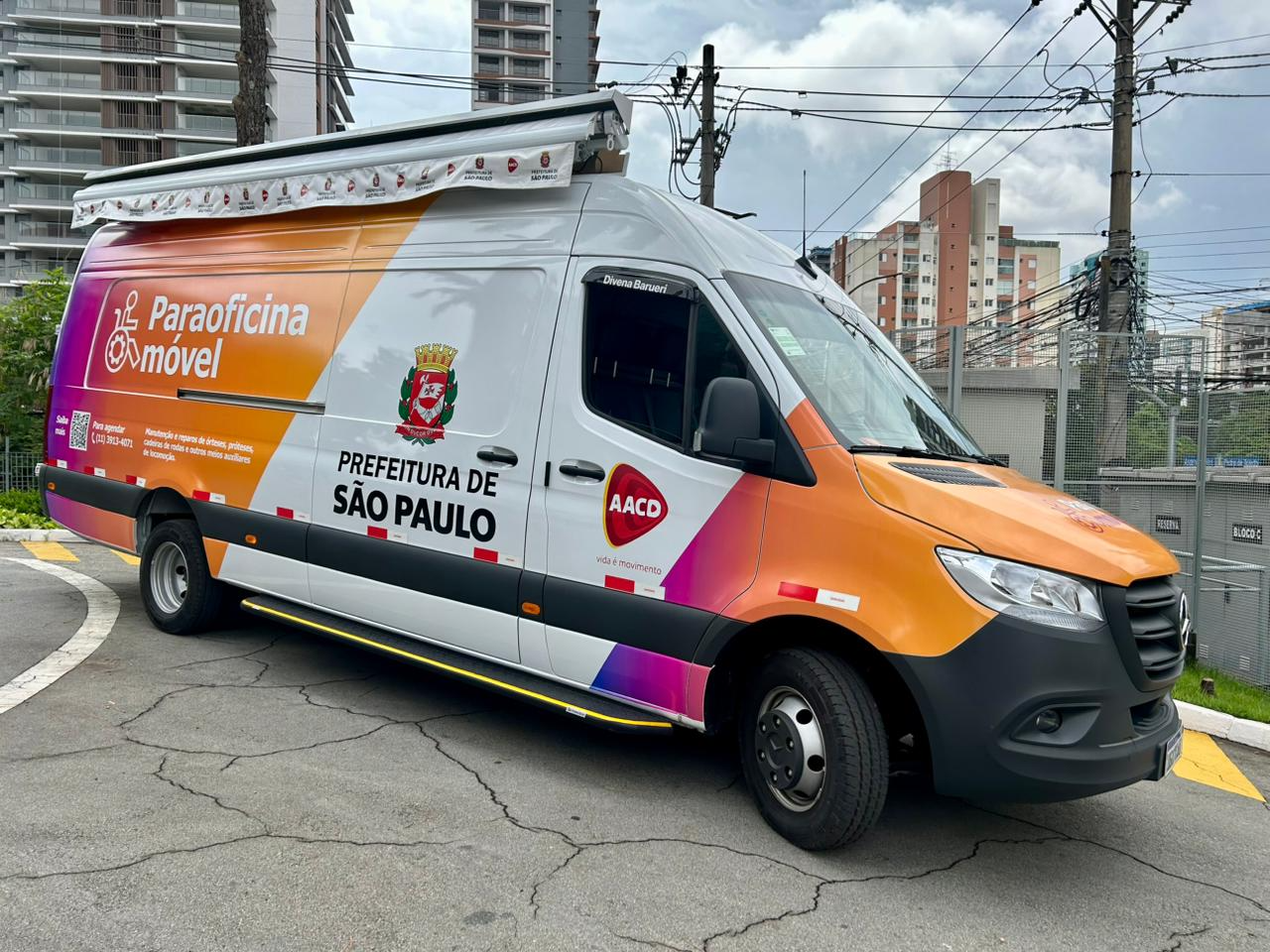 an da Paraoficina Móvel estacionada em área urbana da capital paulista. O veículo, identificado com as marcas da Prefeitura de São Paulo e da AACD, possui estrutura adaptada para atendimento itinerante e é utilizado para oferecer manutenção e pequenos reparos em cadeiras de rodas, órteses, próteses e dispositivos auxiliares de locomoção. Ao fundo, aparecem prédios residenciais e a rede elétrica da cidade.
