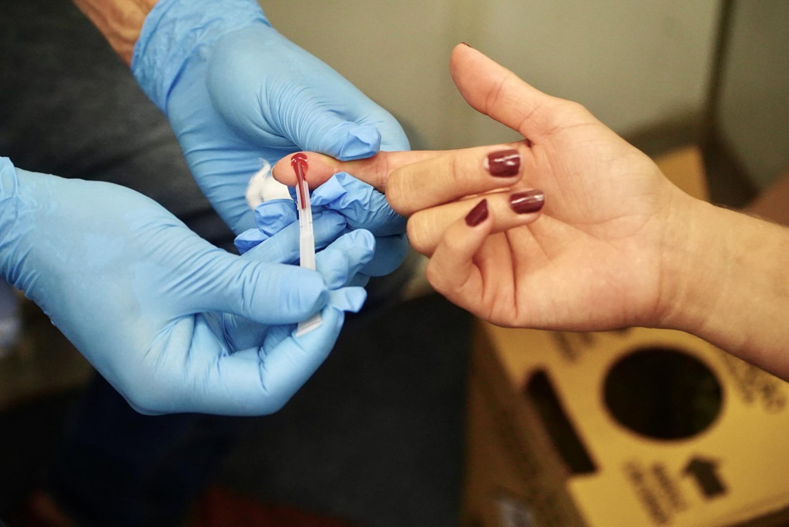 A imagem mostra o momento da coleta de sangue por punção digital para realização de teste rápido. Um profissional de saúde, utilizando luvas azuis, segura o dedo de uma paciente enquanto coleta uma gota de sangue com um dispositivo específico para exame. A cena remete às ações de diagnóstico e prevenção de infecções sexualmente transmissíveis realizadas na rede municipal de saúde.