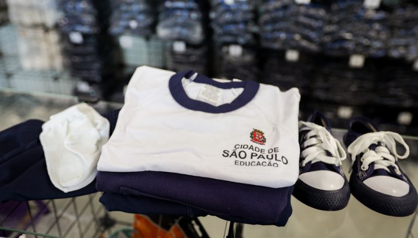 A imagem mostra um conjunto de uniforme escolar organizado sobre uma superfície, possivelmente uma mesa ou carrinho. No centro, há uma camiseta branca dobrada, com gola azul-escura. No lado esquerdo do peito da camiseta, aparece um brasão colorido e o texto “Cidade de São Paulo – Educação”, indicando que é um uniforme da rede municipal de ensino. Abaixo da camiseta, vê-se uma peça de roupa azul-escura dobrada, provavelmente uma calça ou bermuda do uniforme. À esquerda do conjunto, há um par de meias brancas dobradas. À direita, aparece um par de tênis infantis nas cores azul-marinho e branco, com cadarços brancos e biqueiras claras.