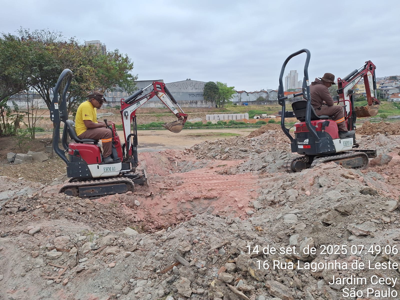 Dois homens estão operando pequenas escavadeiras em um terreno aberto cheio de entulho e terra.Cada um está sentado em sua máquina vermelha, movimentando o braço mecânico para cavar o solo. Ao fundo, aparecem algumas árvores, blocos de concreto empilhados, casas e prédios mais distantes. O céu está nublado, dando um tom acinzentado à cena. No canto inferior direito da imagem há uma marca d’água com data, horário e localização: “14 de set. de 2025, 07:49:06, 16 Rua Lagoinha de Leste, Jardim Cecy, São Paulo”.