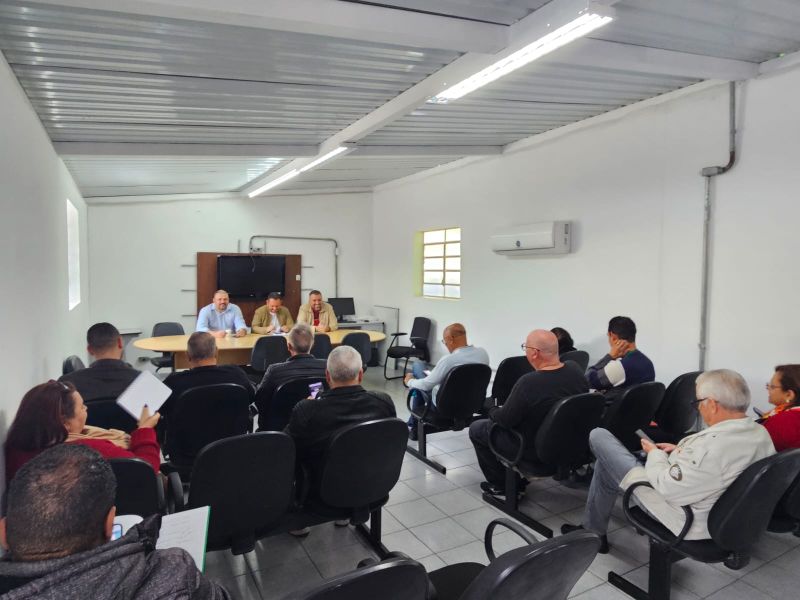 A imagem mostra uma reunião em uma sala simples. Três homens estão sentados à mesa conduzindo a conversa, enquanto cerca de dez pessoas os escutam sentadas em cadeiras.