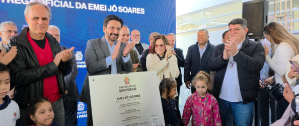 Na imagem, autoridades e convidados participam da inauguração oficial da EMEI José Eugênio Soares (Jô Soares). Em primeiro plano, há uma placa comemorativa da inauguração sendo apresentada. Atrás da placa, várias pessoas aplaudem e sorriem, entre elas o prefeito Ricardo Nunes (de terno cinza e camisa azul clara), acompanhado por secretários municipais e o subprefeito do Itaim Paulista. Também estão presentes crianças em uniforme escolar e familiares, que acompanham o evento ao lado dos representantes da Prefeitura. Ao fundo, há um painel azul com o texto “Entrega Oficial da EMEI Jô Soares