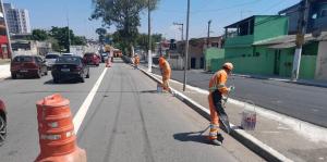 A imagem mostra uma rua de asfalto com duas faixas de rolamento em cada sentido, separadas por uma linha branca contínua. As faixas estão sendo pintadas com uma nova demarcação, com uma linha branca mais larga sendo aplicada na borda da via. Na via, há vários cones de sinalização laranja, indicando a área de trabalho e alertando os motoristas. À direita da via, há uma calçada e, mais ao fundo, algumas casas e prédios de diferentes cores e alturas, incluindo uma construção verde. Três trabalhadores, vestidos com uniformes laranja e com capacetes, estão trabalhando na pintura da via. Um deles está mais próximo da câmera, agachado e pintando a linha branca com um rolo. Outro trabalhador está mais ao fundo, também pintando a via. Um terceiro trabalhador está mais distante, aparentemente preparando a tinta ou outra tarefa relacionada. Há alguns carros trafegando na via, indicando que a obra está sendo realizada com o fluxo de veículos. O céu está azul e ensolarado, sugerindo que a foto foi tirada durante o dia.