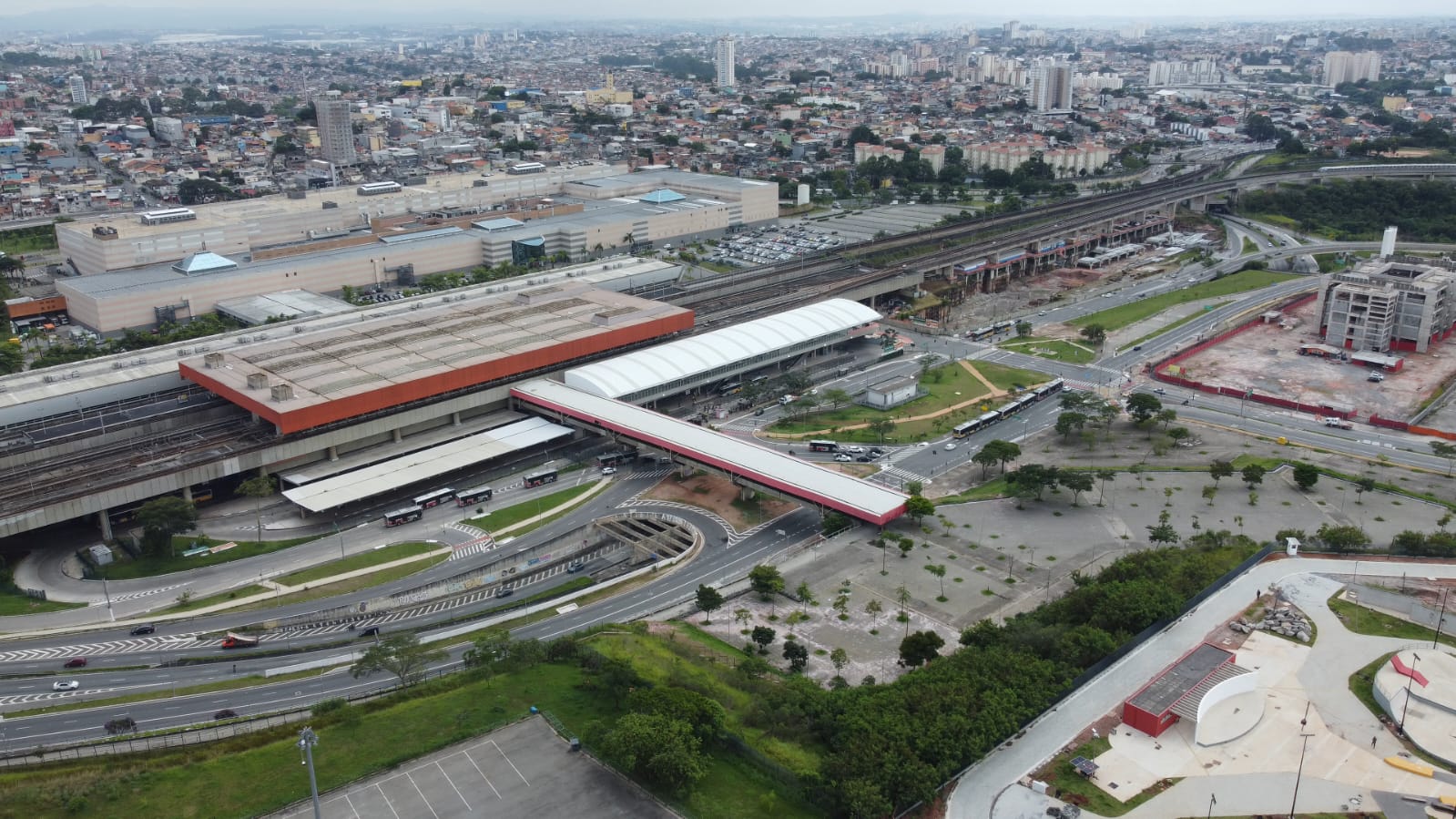 A imagem é uma vista aérea de uma grande estação de transporte público, provavelmente uma estação de trem ou metrô, localizada em uma área urbana densamente povoada. No centro da imagem, destacam-se várias plataformas cobertas com telhados de cor clara, possivelmente brancos ou cinza claro, com estruturas de apoio em tons de laranja ou vermelho. As plataformas estão posicionadas sobre trilhos de trem, que se estendem em diferentes direções, indicando uma importante linha de transporte. Há várias vias de acesso e áreas de circulação ao redor da estação, incluindo viadutos e ruas com fluxo de veículos. É possível observar automóveis e ônibus em movimento. Ao redor da estação, há uma vasta área com edifícios de diferentes tamanhos e alturas, indicando uma cidade grande. Há também áreas verdes, como gramados e algumas árvores, mas a predominância é de construções urbanas. No canto inferior direito, há uma estrutura que parece ser um parque ou área de lazer, com algumas formas geométricas e áreas gramadas. A imagem sugere uma infraestrutura de transporte público moderna e integrada à área urbana, com uma grande movimentação de pessoas e veículos.