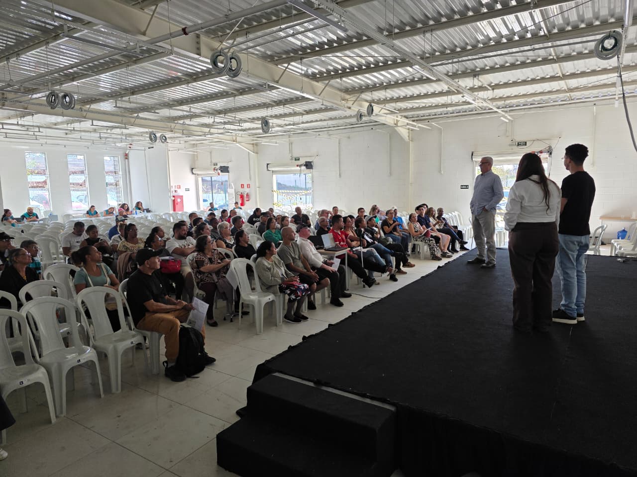 Pessoas acompanham o chamamento para escolha de seus apartamentos em um auditório, enquanto três participantes falam no palco à frente do público.