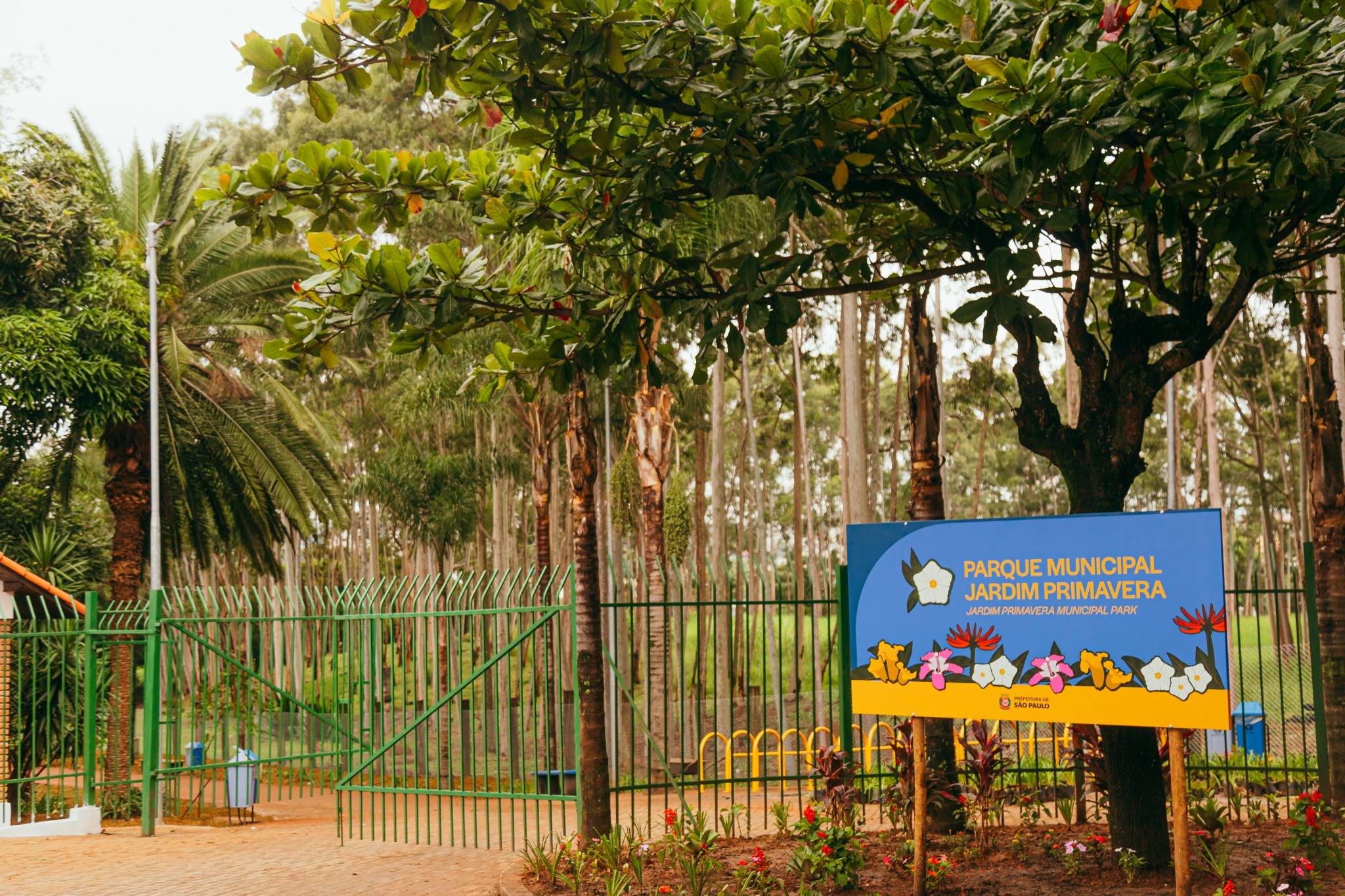 Imagem de entrada de um parque arborizado, com predominância de tons verdes e terrosos. Em primeiro plano, há um portão metálico pintado de verde, parcialmente aberto, com grade vertical e estrutura simples. Ao lado direito, destaca-se uma placa retangular em azul com detalhes em amarelo, onde está escrito “Parque Municipal Jardim Primavera”, acompanhada de ilustrações de flores coloridas em tons de branco, amarelo, rosa e vermelho. A placa está fixada em dois suportes de madeira e cercada por um pequeno jardim com flores vermelhas e folhagens verdes. Acima, uma árvore de copa larga com folhas verdes densas cria sombra sobre a placa. Ao fundo, há diversas árvores altas e finas, formando uma paisagem de mata, além de um poste de iluminação branco à esquerda. O chão próximo à entrada é de piso claro, em tom bege, contrastando com a vegetação ao redor.