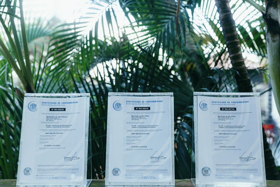 Fotografia de três certificados da Associação Brasileira de Normas Técnicas (ABNT) expostos lado a lado sobre uma superfície de madeira, em ambiente externo. Os documentos estão acomodados em suportes transparentes e representam certificações concedidas ao Município de São Paulo. Ao fundo, uma densa vegetação tropical com palmeiras e folhagens verdes cria um cenário natural, reforçando a relação da premiação com sustentabilidade e meio ambiente. A iluminação natural destaca os certificados em primeiro plano, enquanto a vegetação aparece levemente desfocada ao fundo.