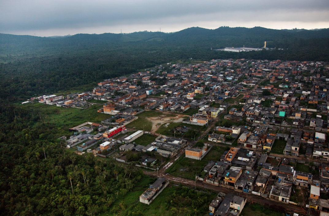 #ParaCegoVer  Vista aérea que mostra o contraste entre a natureza e a expansão urbana. À esquerda, há uma densa floresta verde-escuro. À direita, estende-se uma comunidade periférica com casas baixas, muitas de tijolos aparentes ou lajes, organizadas ao longo de ruas estreitas. Ao fundo, morros cobertos por vegetação tocam um céu nublado e cinzento, destacando-se uma grande estrutura branca com uma torre amarela no topo de uma das colinas.