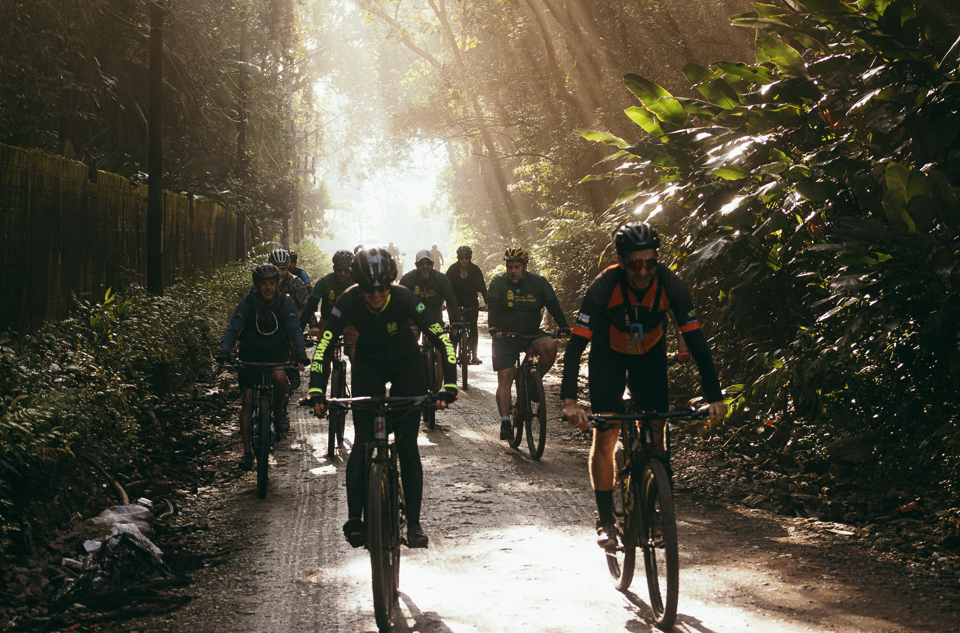 Fotografia mostra um grupo de ciclistas pedalando por uma estrada de terra cercada por vegetação densa da Mata Atlântica. Ao todo, cerca de dez pessoas seguem em direção à câmera, todas utilizando capacete e bicicletas de diferentes modelos. A luz do sol atravessa as copas das árvores, criando feixes luminosos que iluminam o caminho e reforçam a atmosfera de contato com a natureza. Nas laterais da estrada, há árvores altas, arbustos e folhagens verdes, compondo um cenário de floresta preservada.