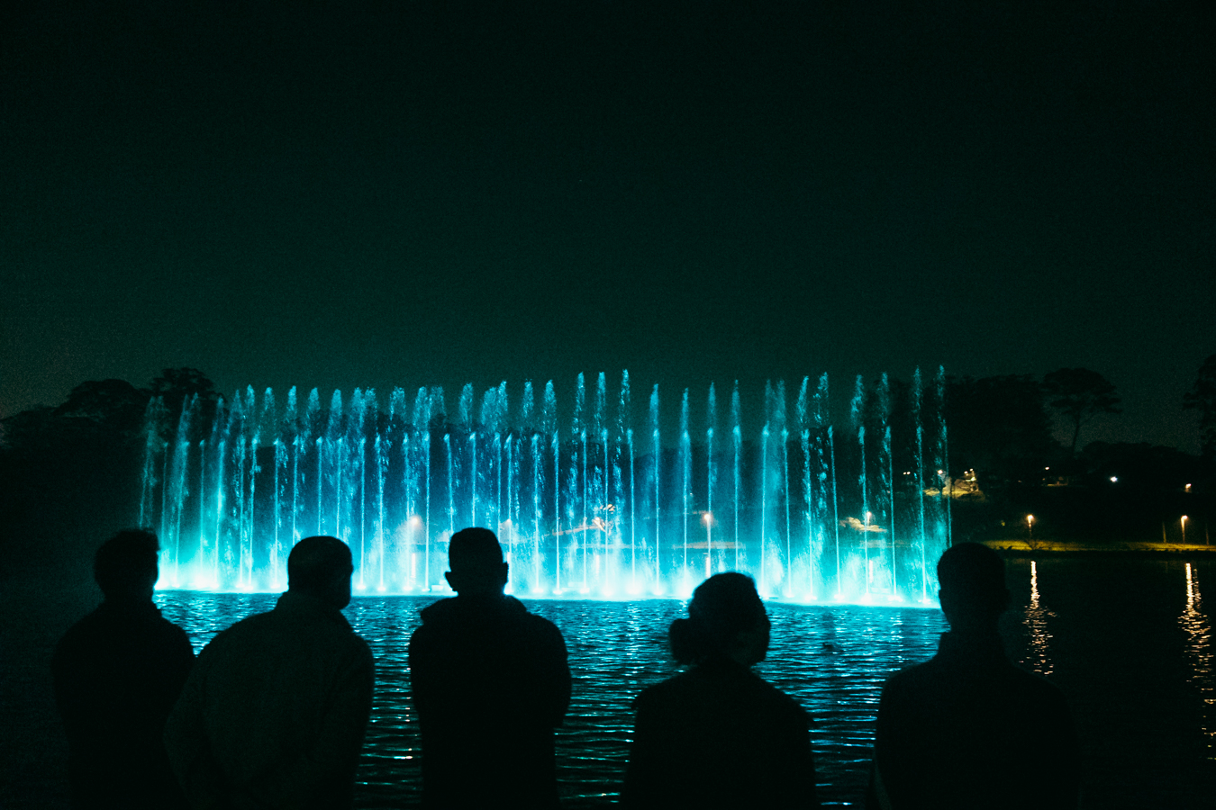 A cena captura uma performance impressionante de uma fonte musical em um lago, sob o manto de uma noite sem lua, onde apenas o azul elétrico das águas rompe a escuridão. O enquadramento é horizontal e a imagem tem um grão cinematográfico sutil.
