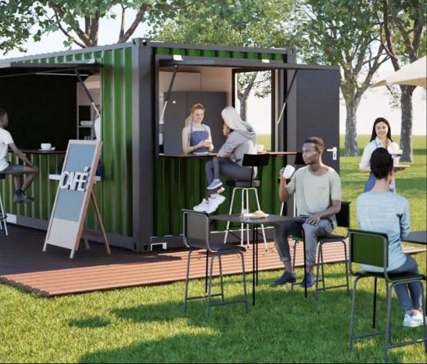 Imagem mostra um quiosque de café feito a partir de um contêiner metálico verde, instalado em uma área gramada de parque. O contêiner está aberto na lateral, formando um balcão onde uma atendente serve bebidas. À frente, há mesas e bancos altos, também em tons de verde e preto. Pessoas adultas estão sentadas e em pé, conversando e tomando café. Ao fundo, aparecem árvores com copa verde, indicando um ambiente ao ar livre, iluminado pela luz do dia.