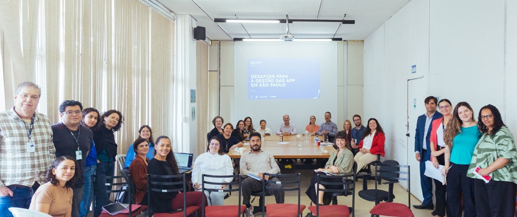 Fotografia mostra cerca de 20 pessoas reunidas em uma sala de reuniões, posicionadas ao redor de uma mesa retangular e olhando para a câmera. Ao fundo, uma projeção exibe o título "Desafios para a gestão das APP em São Paulo". O ambiente é amplo, iluminado por janelas laterais com persianas, transmitindo um clima de encontro técnico e colaborativo.