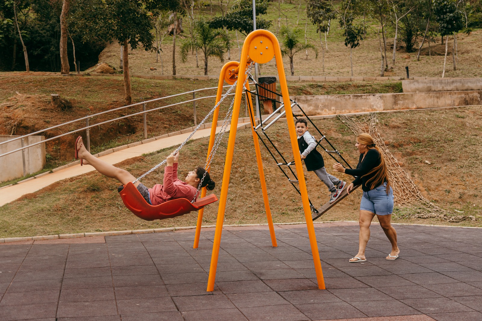 Fotografia de um playground em um parque municipal. Em primeiro plano, uma criança se diverte em um balanço adaptado com assento em formato de concha, enquanto outra criança sobe em uma estrutura de cordas integrada ao brinquedo. Ao lado, uma mulher acompanha e incentiva a brincadeira. O piso é emborrachado e, ao fundo, há um gramado com árvores, uma rampa acessível com corrimão e um cenário de área verde, reforçando o ambiente de lazer e contato com a natureza.