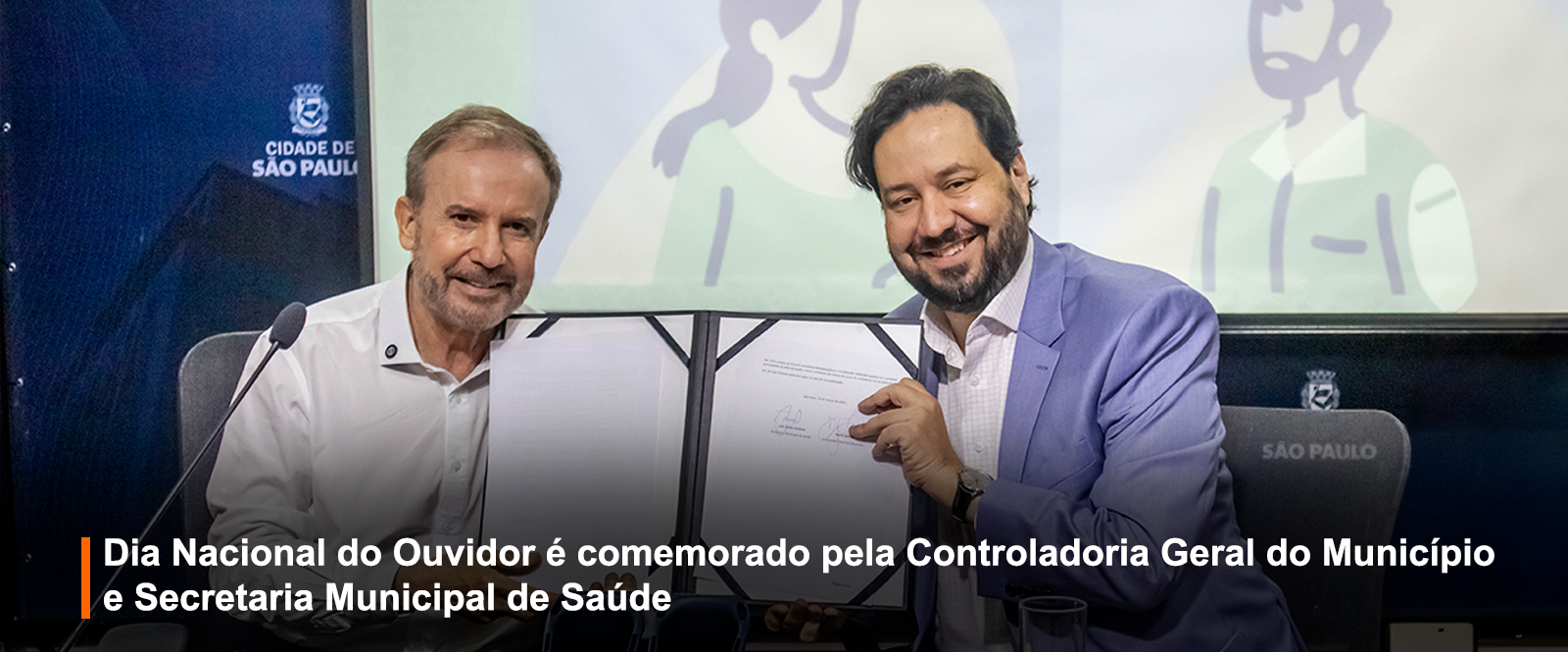 Fotografia de evento institucional mostra dois homens sorrindo, segurando um documento. Um veste camisa branca, o outro blazer azul claro. Ao fundo, tela com ilustrações e o brasão da Cidade de São Paulo. Faixa preta inferior: 