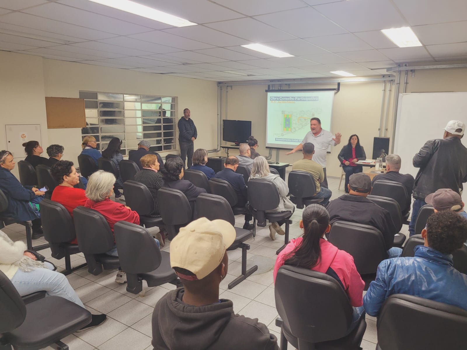 Foto da reunião tirada de trás, com pessoas sentadas e o Subprefeito na frente 