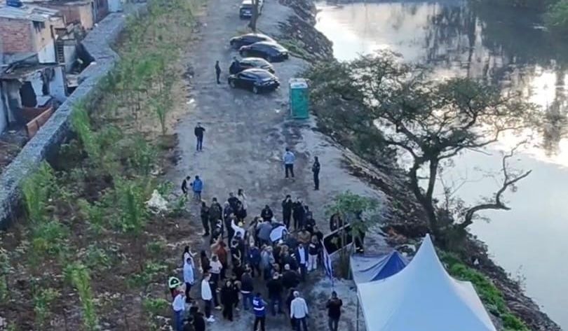 Área às margens do Rio Tietê recebe evento relacionado ao projeto de recuperação do Pantanal. Na imagem aérea, é possível observar um grupo de pessoas reunidas próximo ao rio, além de uma tenda de apoio, veículos e uma área com vegetação em processo de recuperação. Ao fundo, aparecem o curso d'água, árvores, um muro de gabião e moradias da região.