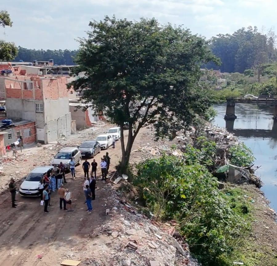 A imagem mostra a poluição presente às margens do Rio Tietê, funcionários públicos trabalhando e moradias localizadas em área de alto risco. 