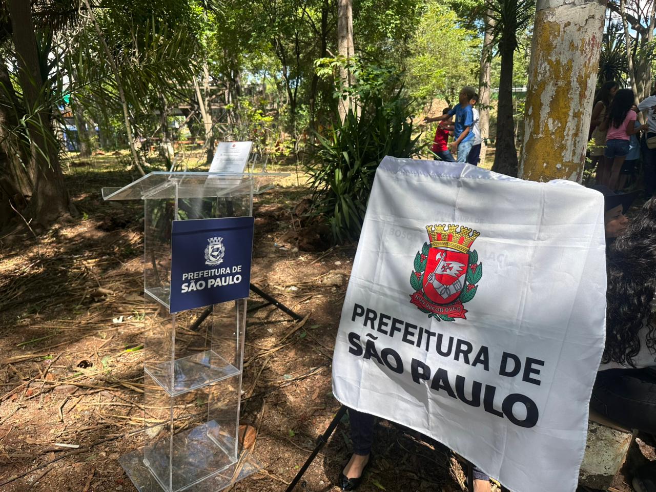 A sequência de fotos registra um evento de plantio e inauguração da Prefeitura de São Paulo em uma área arborizada. Na primeira imagem, um trabalhador com uniforme laranja, colete de sinalização amarelo e capacete azul utiliza uma enxada para preparar a terra ao redor de uma muda de árvore. Na segunda foto, um grupo de pessoas participa do plantio; uma mulher com blusa de estampa de onça e um senhor de camisa listrada manuseiam uma muda, cercados por outros adultos e funcionários da prefeitura em um terreno com folhas secas e luz solar filtrada pelas árvores. A terceira imagem foca nos preparativos oficiais, mostrando um púlpito de acrílico transparente com o logo azul da Prefeitura de São Paulo e, ao lado, uma bandeira branca com o brasão colorido da cidade. Ao fundo das cenas, é possível ver a vegetação densa do local, grades de proteção e crianças brincando, indicando que a ação ocorre em um parque ou bosque público durante um dia ensolarado.