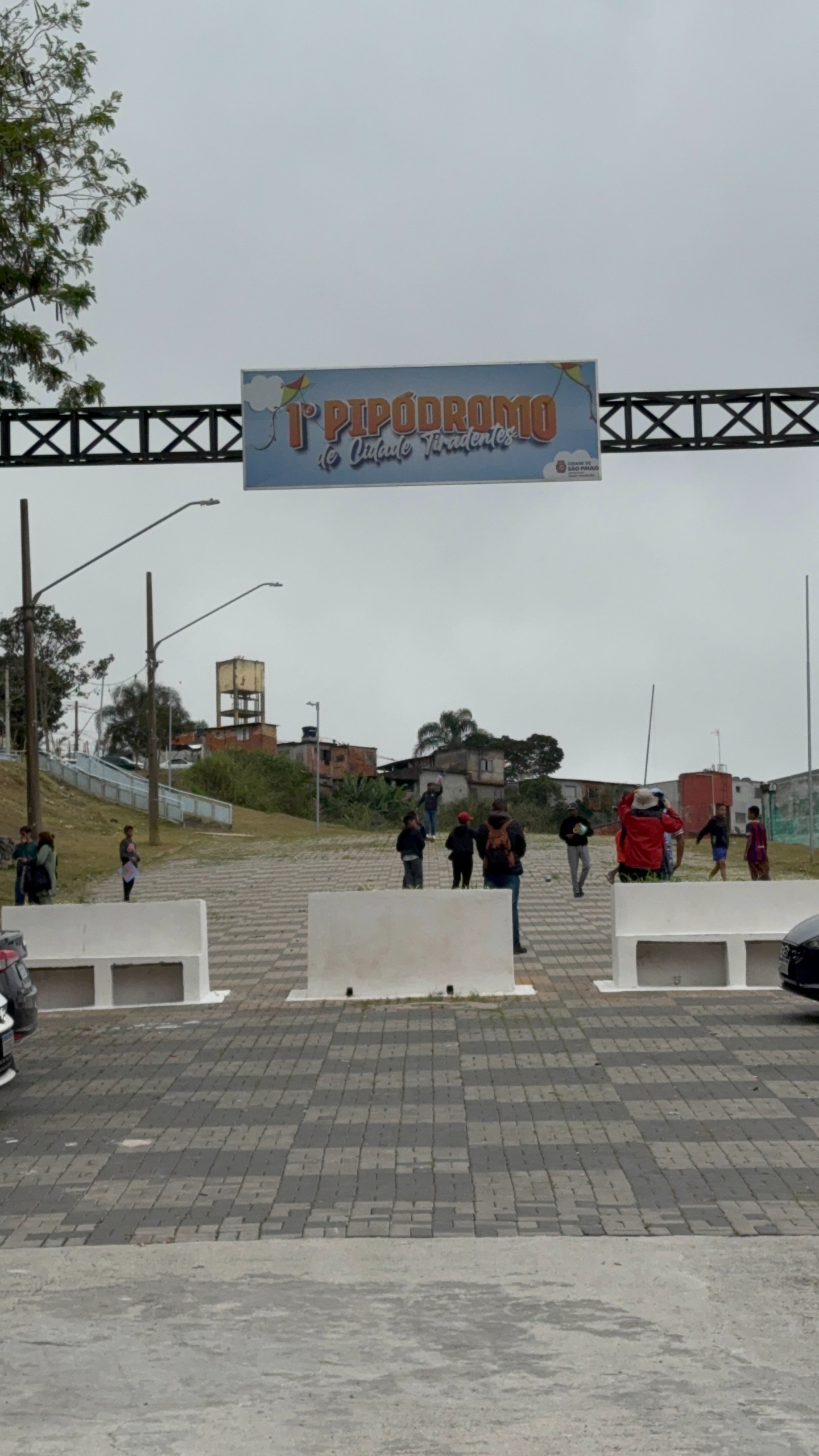 A foto na vertical, tirada ao nível do chão, mostra a entrada de uma praça sob um céu nublado e cinzento. Na parte superior, uma estrutura metálica preta atravessa o espaço de um lado a outro, sustentando ao centro uma placa retangular onde se lê "1º PIPÓDROMO de Cidade Tiradentes", com ilustrações de pipas nas pontas e a logo da Prefeitura de São Paulo no canto inferior direito. Logo abaixo da estrutura, blocos de concreto brancos delimitam o acesso ao piso pavimentado em blocos cinzas e pretos. No espaço da praça, várias pessoas transitam e conversam. Ao fundo, em terreno elevado, avistam-se postes de iluminação pública, árvores, caixas-d'água e construções residenciais da comunidade local.